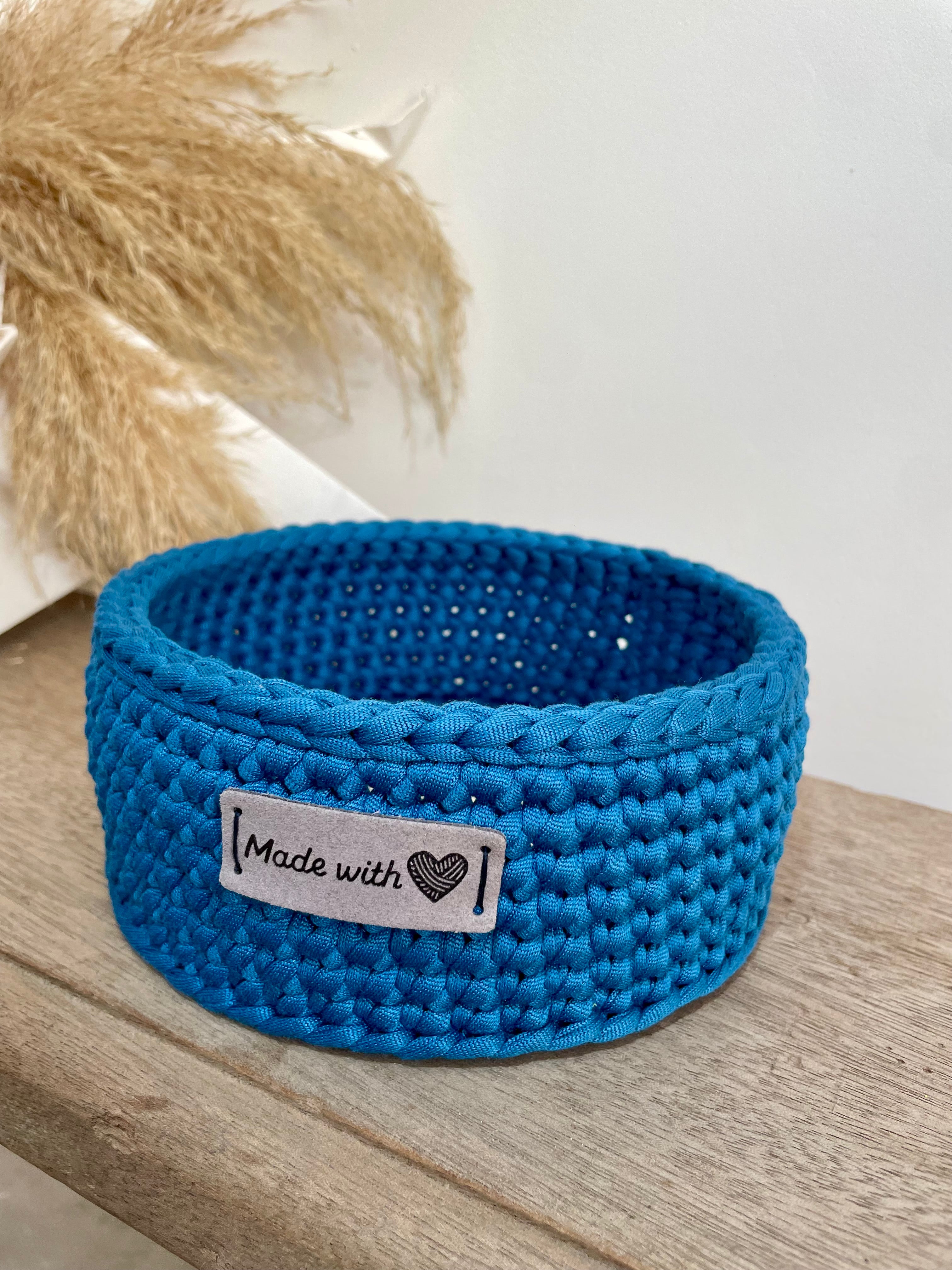 Panière bleue crochetée artisanalement sur laquelle est cousue une étiquette en cuir beige made with heart. La panière est posée sur un banc en bois à coté d'une décoration en papier et fleur de la pampa