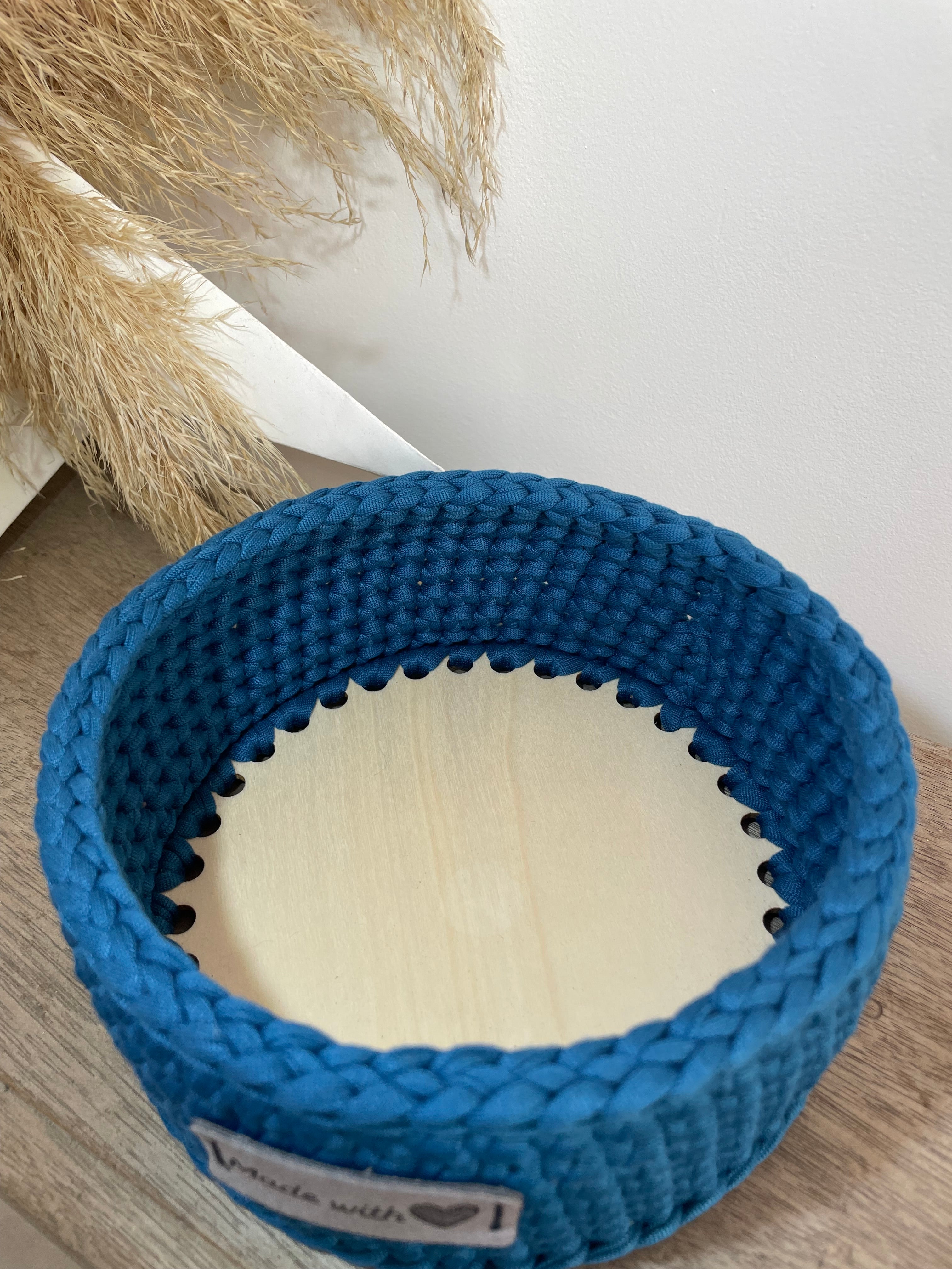 Panière bleue avec un fond en bois crochetée artisanalement. À coté est posée une décoration en papier et fleur de la pampa