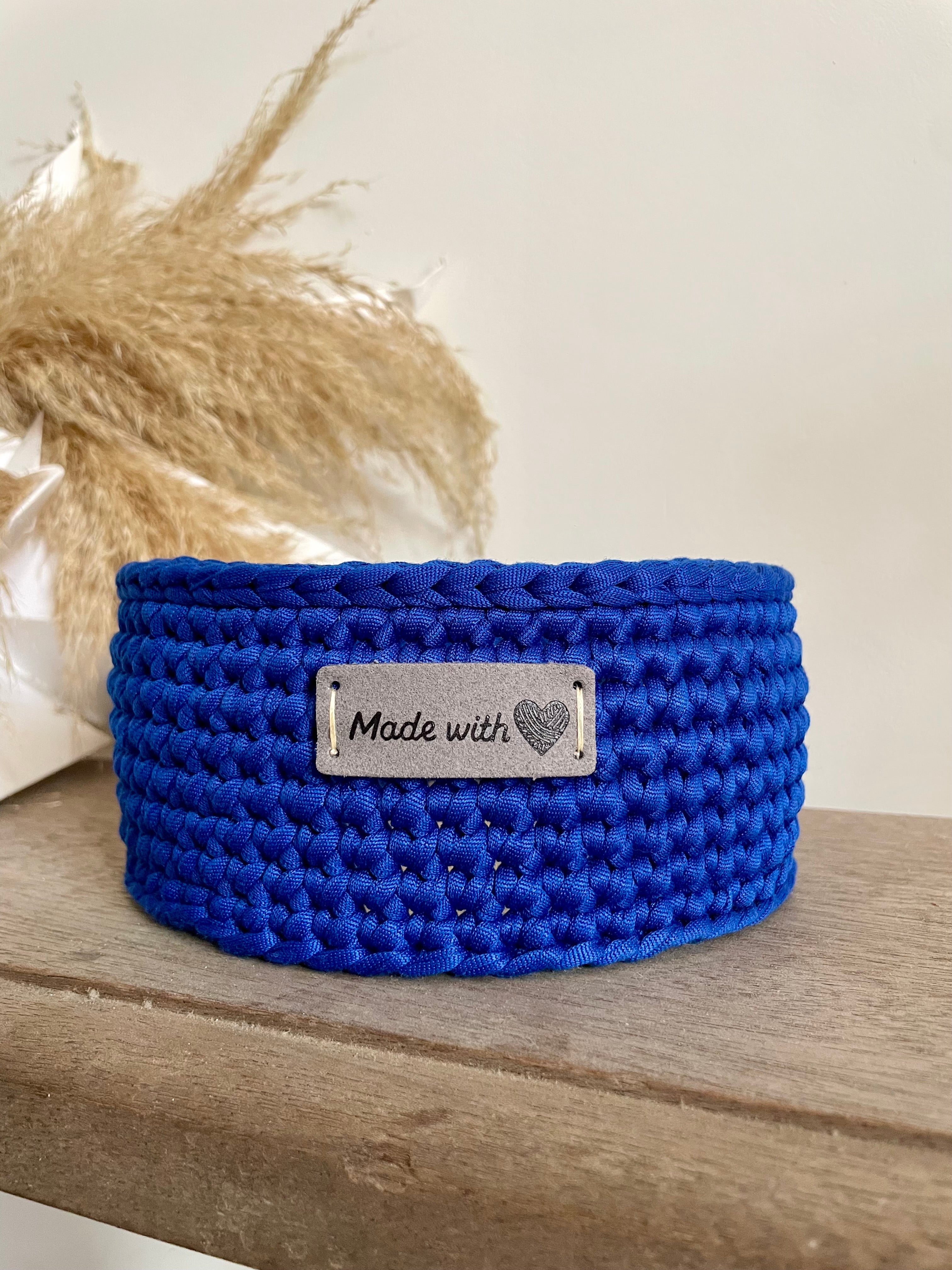 Paniere bleue fabriquée au crochet avec une étiquette grise en cuir made with love. Panière posée sur un banc en bois à coté d'une décoration en papier et fleur de la pampa