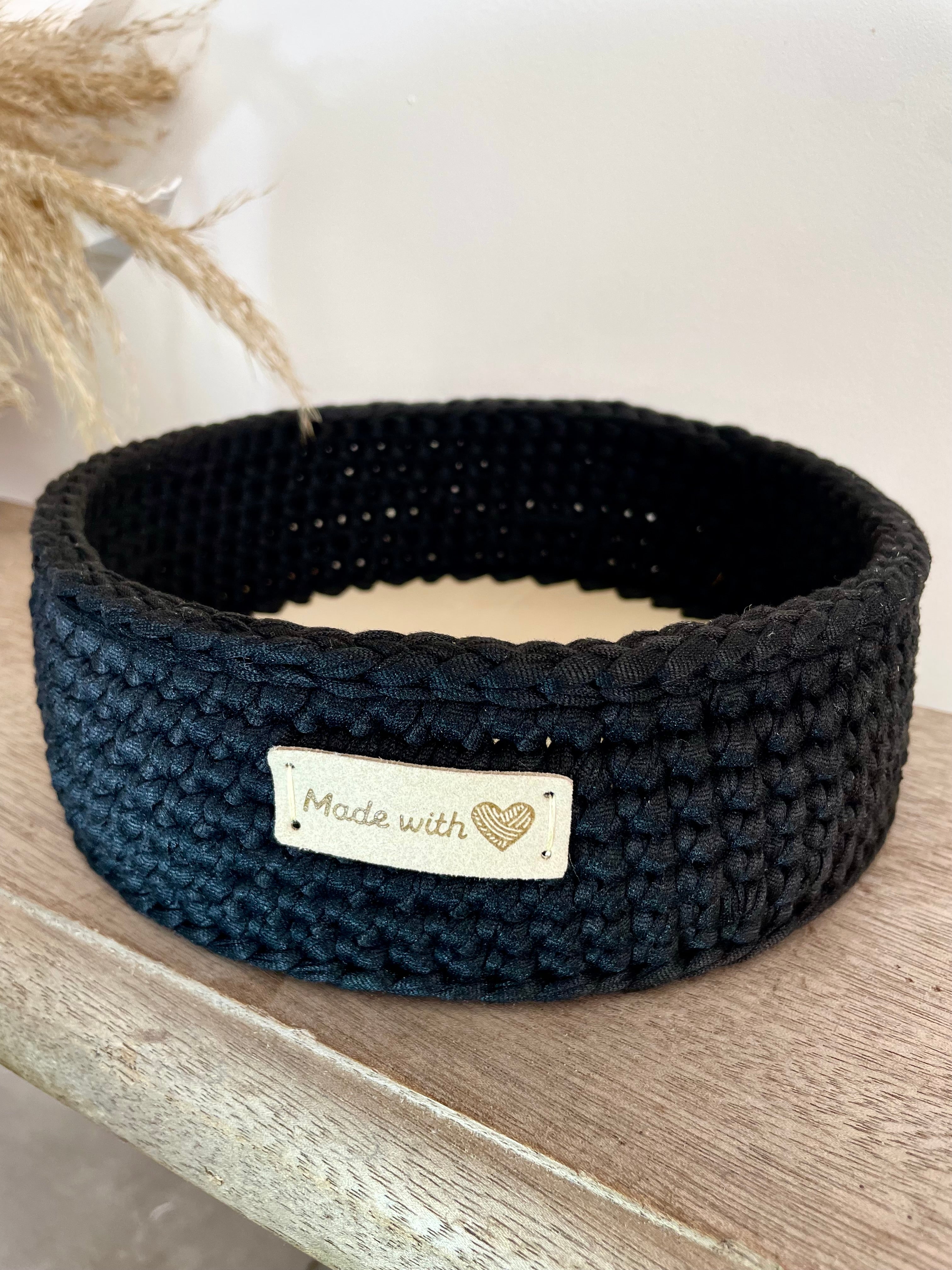 Panière crochetée noire, posée sur un banc en bois.Il y a une étiquette beige en cuir cousue made with love