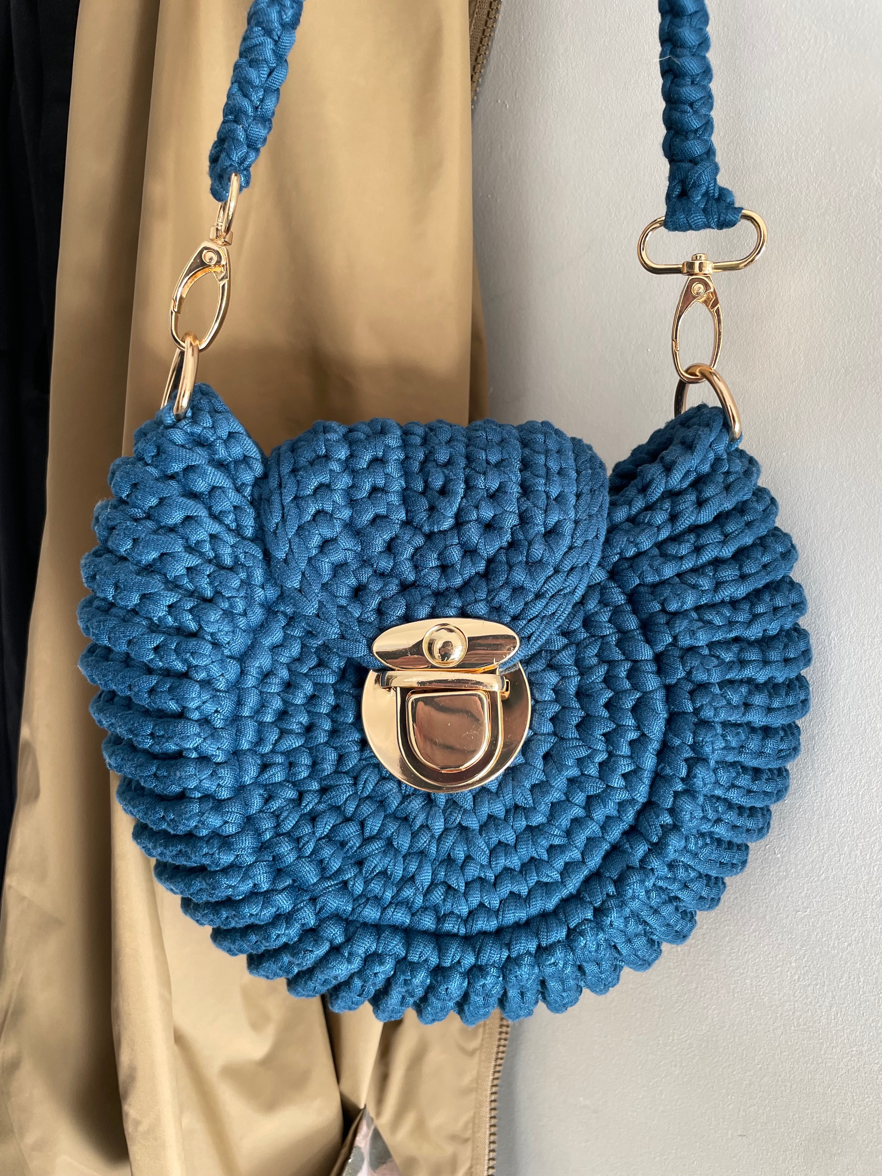 Sac en forme de coquillage bleu avec une attache dorée accroché devant un morceau de veste beige