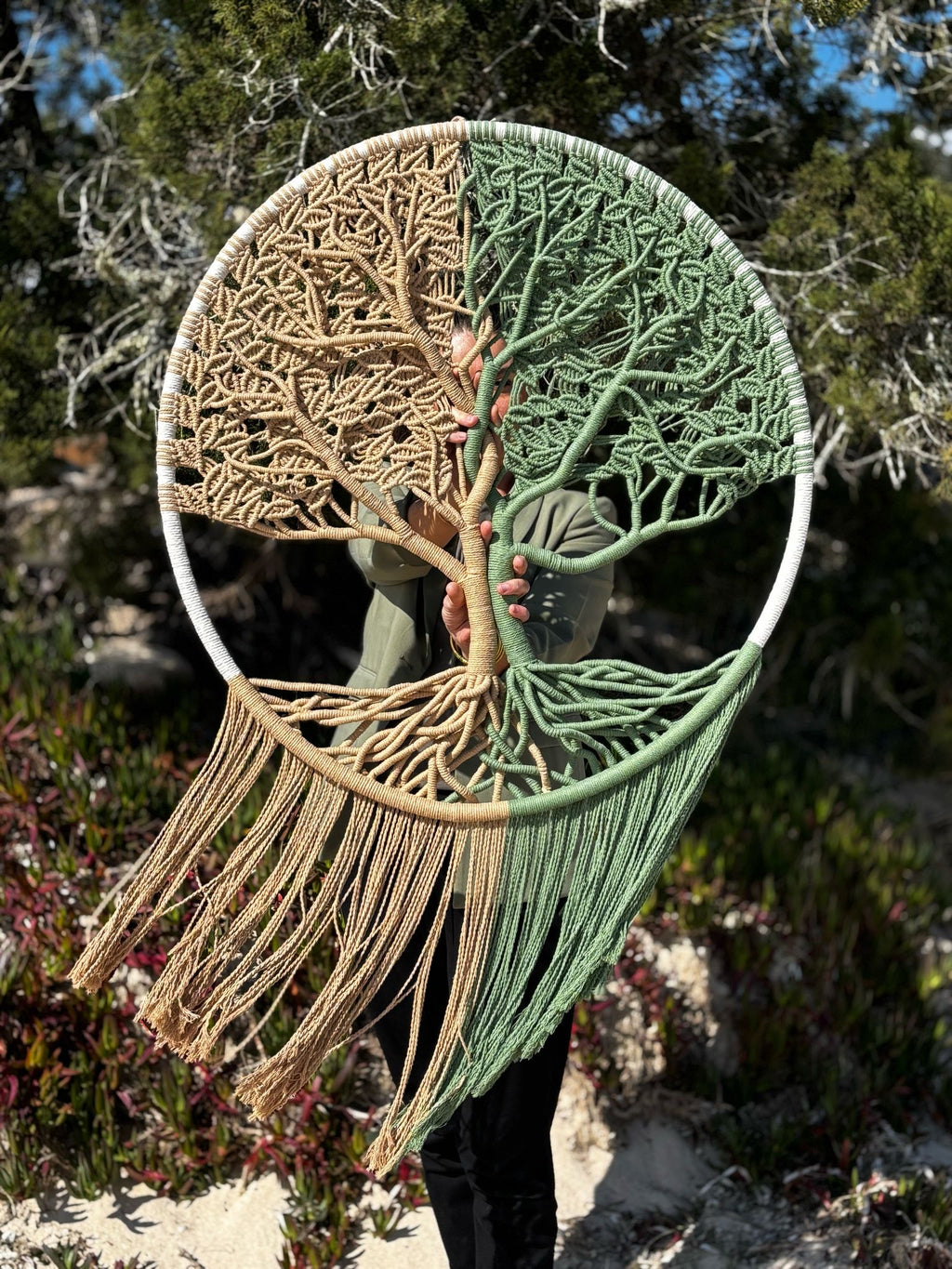 la créatrice se tient debout à la plage avec son arbre de vie en macramé tendu devant elle. l’arbre est de couleur beige marron à gauche et vert à droite 