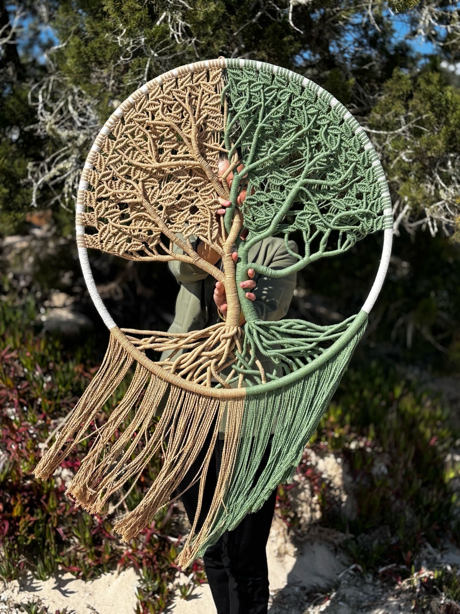 la créatrice se tient debout à la plage avec son arbre de vie en macramé tendu devant elle. l’arbre est de couleur beige marron à gauche et vert à droite 