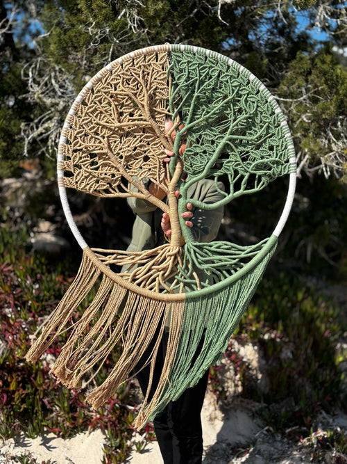 la créatrice se tient debout à la plage avec son arbre de vie en macramé tendu devant elle. l’arbre est de couleur beige marron à gauche et vert à droite 
