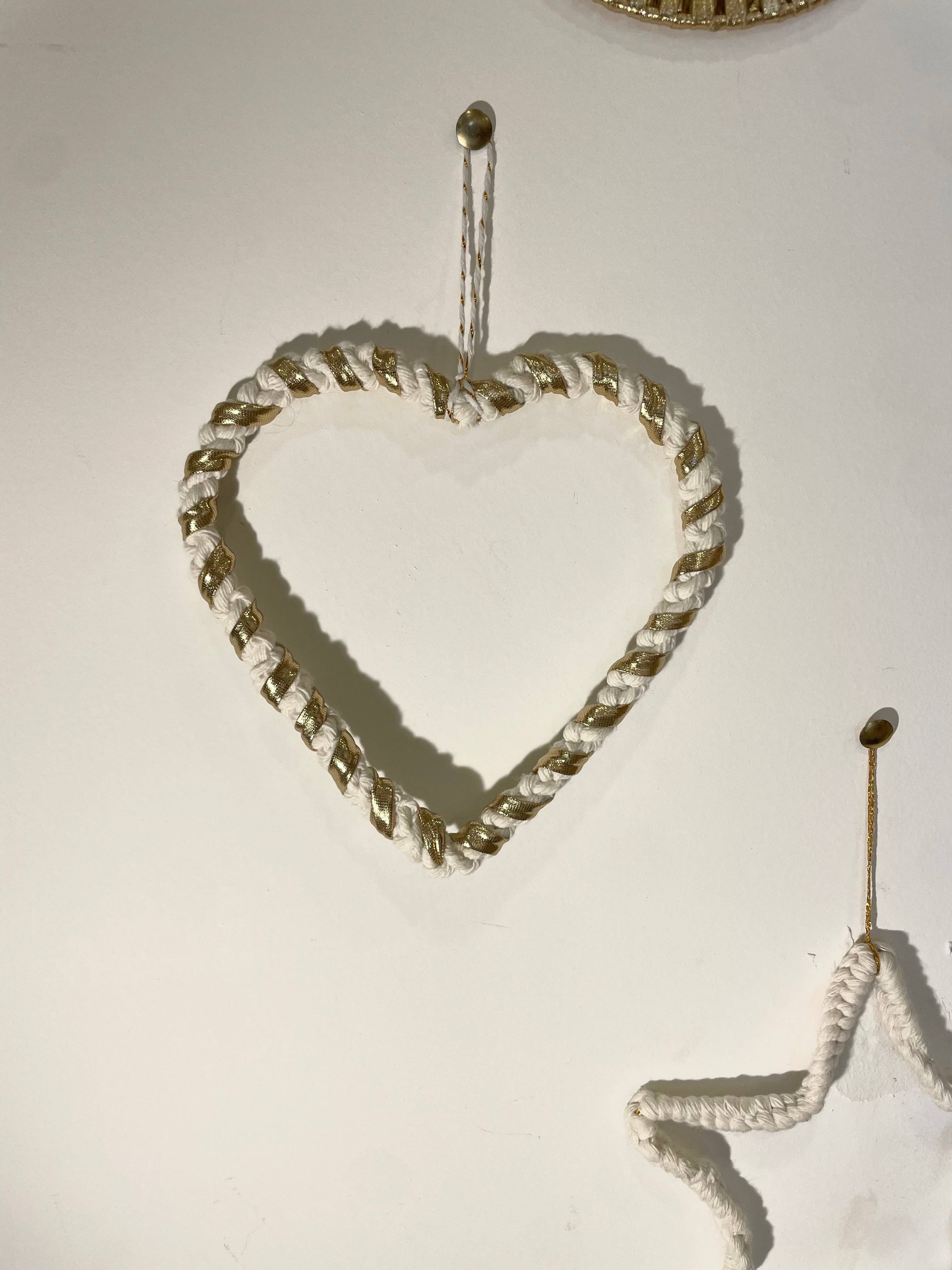 cœur tressé avec la technique macramé suspendu par un fil blanc et doré à un mur