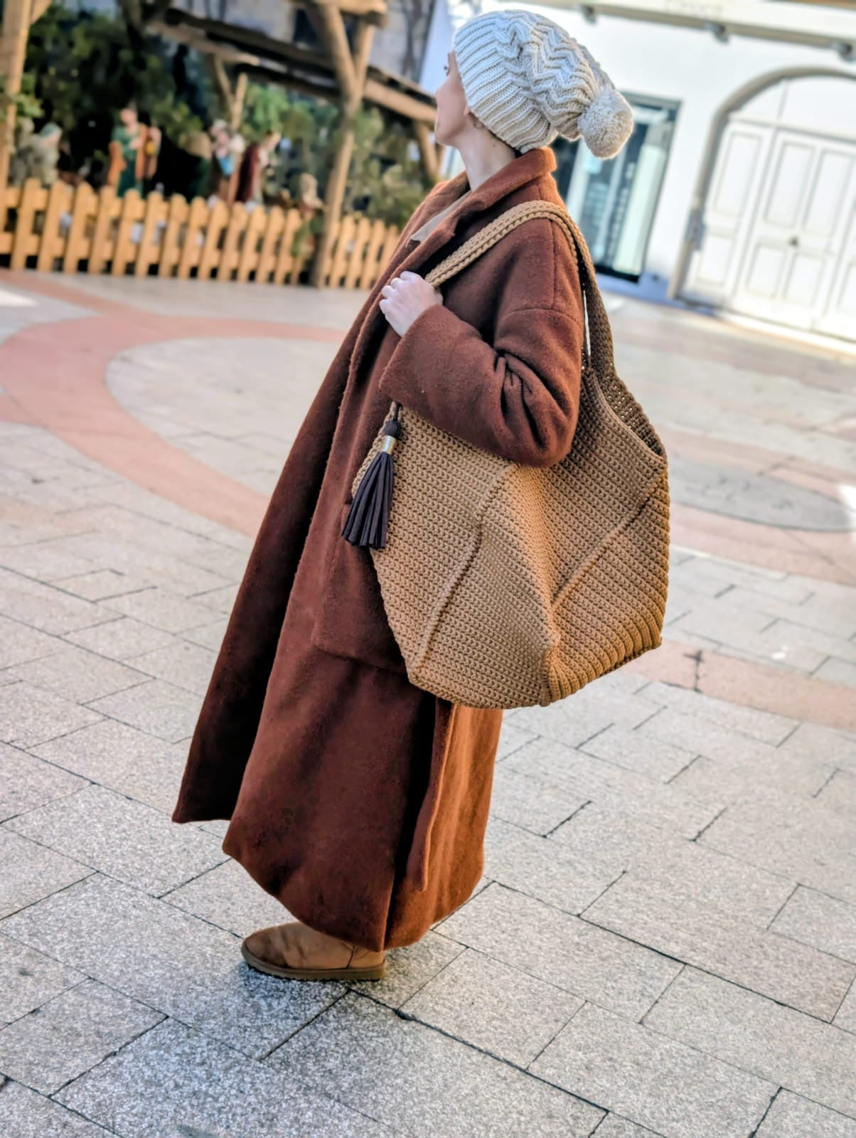 une femme pose dans une rue pavée manteau marron foncé et bonnet beige avec un grand sac cabas couleur caramel et un pompon marron doré accroché à une des anses