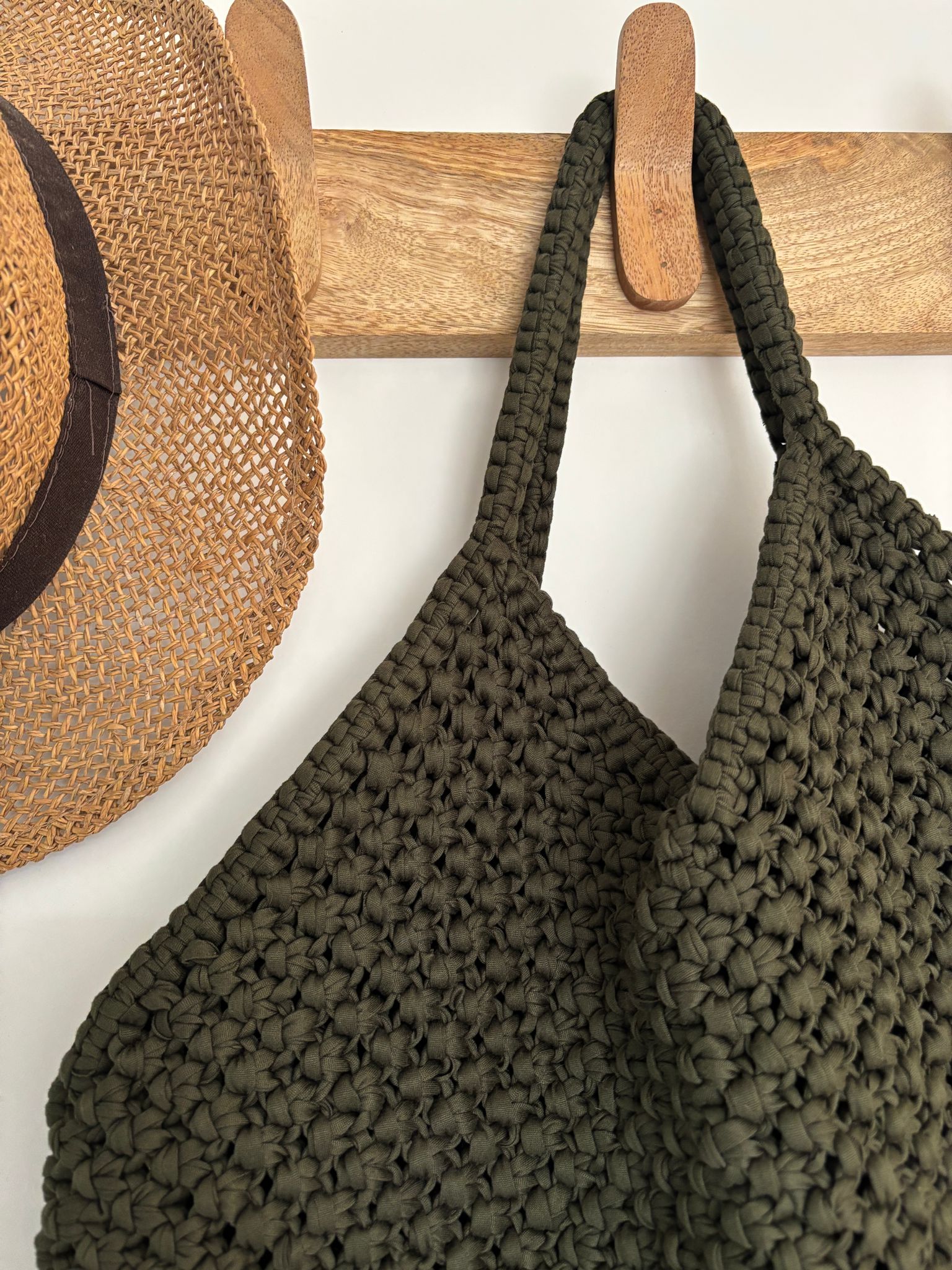 sac de forme triangulaire fabriqué en macramé couleur kaki accroché sur une patère à coté d'un chapeau de paille 
