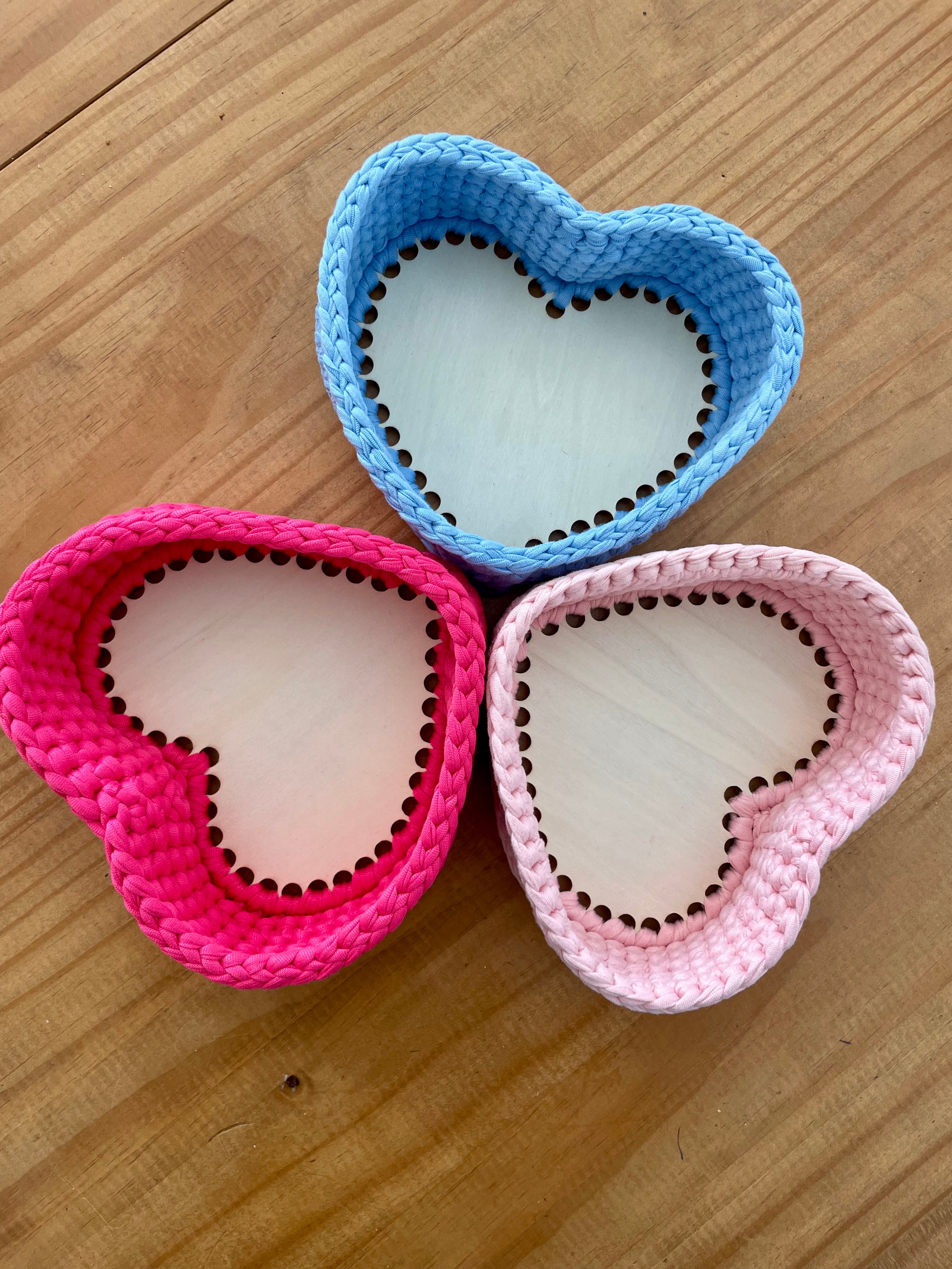 trois panières fabriquées au crochet en forme de cœur de couleurs différentes rose pâle, rose fuchsia et bleu clair avec un fond en bois